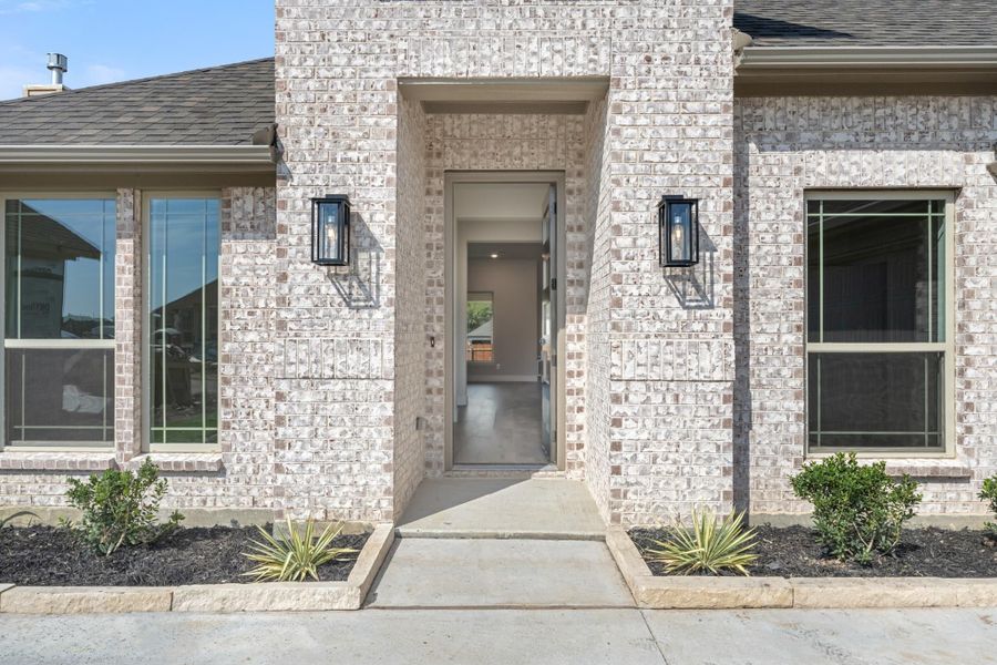 Front entry of a brick home with an open door, two black wall sconces, and a concrete walkway with landscaping.