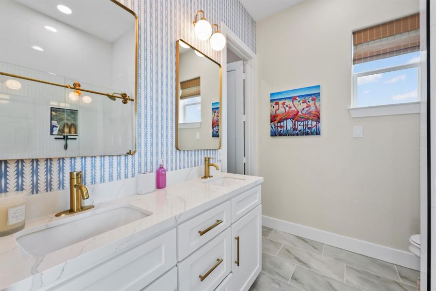 This bathroom features a double vanity with elegant gold fixtures, a stylish blue and white patterned Serena & Lily wallpapered wall, and modern lighting. A window provides natural light, complemented by a vibrant flamingo artwork, creating a bright and inviting space.