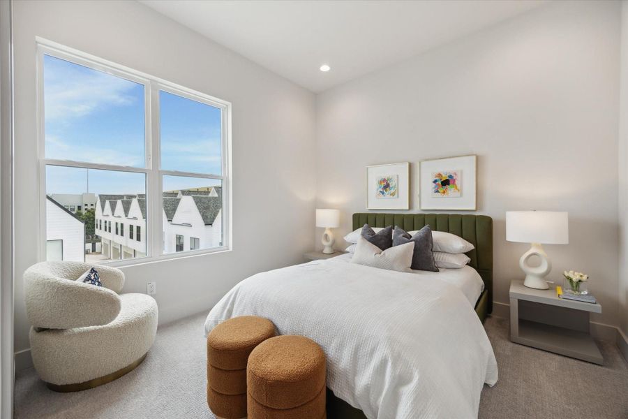 This photo showcases a bright, modern bedroom with a large window offering a view of neighboring houses. The room features a comfortable bed with an upholstered headboard, stylish lamps, and tasteful artwork. Cozy seating and neutral tones enhance the inviting atmosphere.