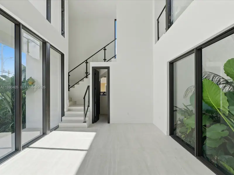 Double-height ceilings, floor-to-ceiling windows with black metal frames, stone-finish flooring, and a staircase featuring glass railing panels with black metal handrails Double-height ceilings, floor-to-ceiling windows with black metal frames, stone-finish flooring, and a staircase featuring glass railing panels with black metal handrails