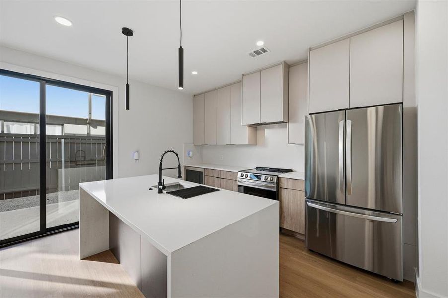 Kitchen features recessed lighting and pendant lighting over the island. Light wood cabinetry extends throughout with built-in microwave and stainless steel refrigerator. Quartz waterfall island complements the gas range and black faucet. Sliding glass door provides backyard access. Vinyl flooring extends throughout.