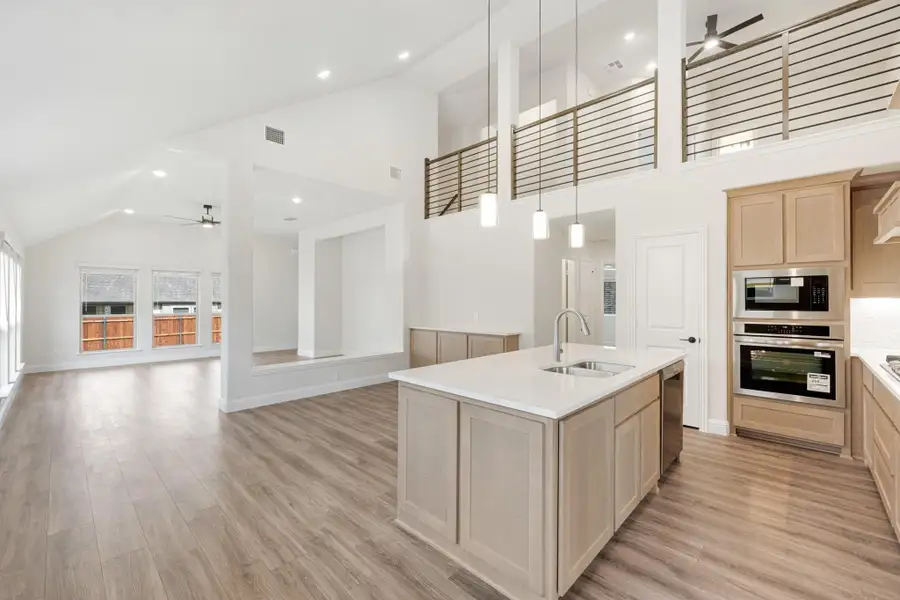 Open-concept kitchen with large island, light wood cabinets, double ovens, and two-story ceiling with upper loft railing.