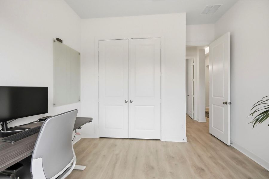Bright and versatile home office/bedroom with sleek laminate flooring, a modern desk setup, and ample storage space behind double doors.