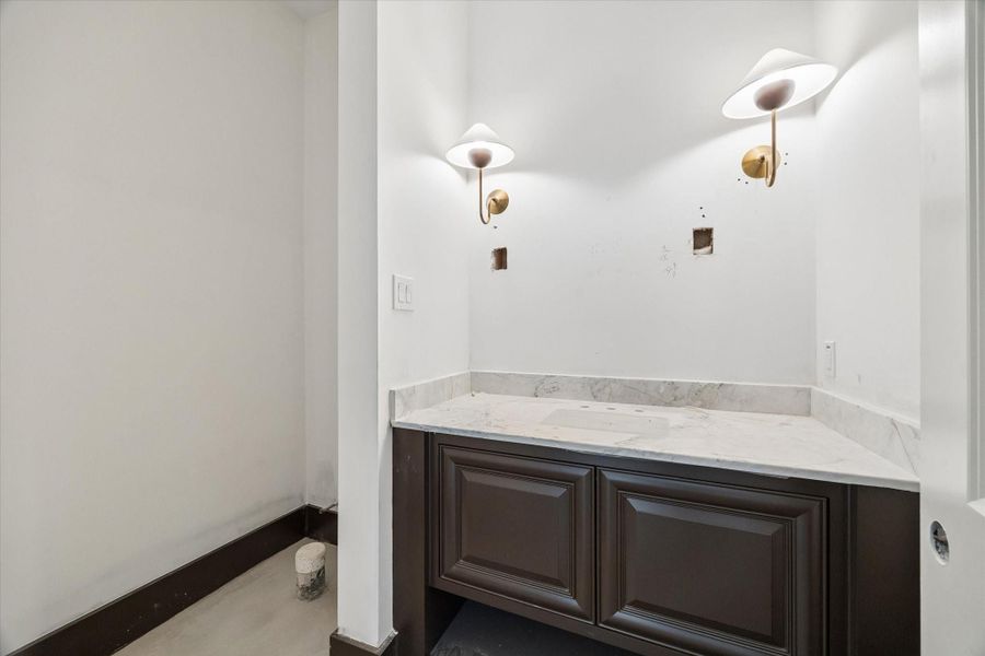Stylish downstairs powder bath featuring a rich furniture style vanity paired with a light stone countertop for a striking contrast. Designer wall sconces add a warm, elevated touch, while the clean lines and neutral palette create a sophisticated space ideal for guests.