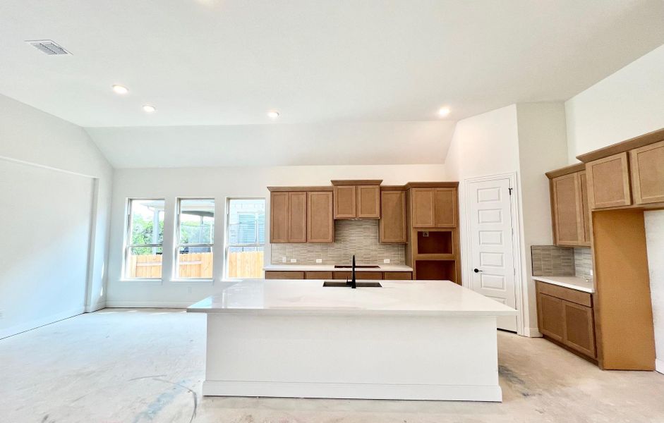 Kitchen with vaulted ceiling, decorative backsplash, brown cabinetry, light stone countertops, and concrete floors Kitchen with vaulted ceiling, decorative backsplash, brown cabinetry, light stone countertops, and concrete floors