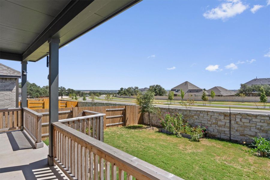 An elevated covered balcony overlooks a grassy backyard enclosed by stone and wood fencing.
