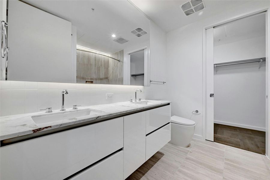 The primary bathroom showcases a dual vanity with sleek white cabinets, providing ample storage & a modern aesthetic plus a large horizontal mirror, enhancing the spacious feel and functionality of the area. The shower and tub combo is designed with floor to ceiling tiles. Additionally, the space includes a walk-in closet, offering convenient storage and organization. Photo is model unit #2406.