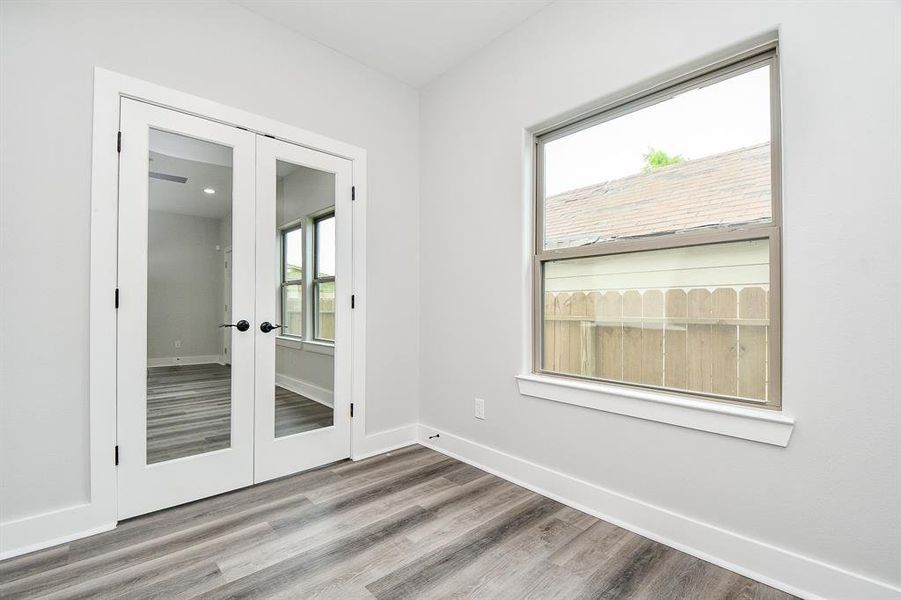 This is a bright, modern in home office or study room featuring gray wood-look flooring, fresh white walls, a large window for ample natural light, and double French doors leading to an adjoining family room.