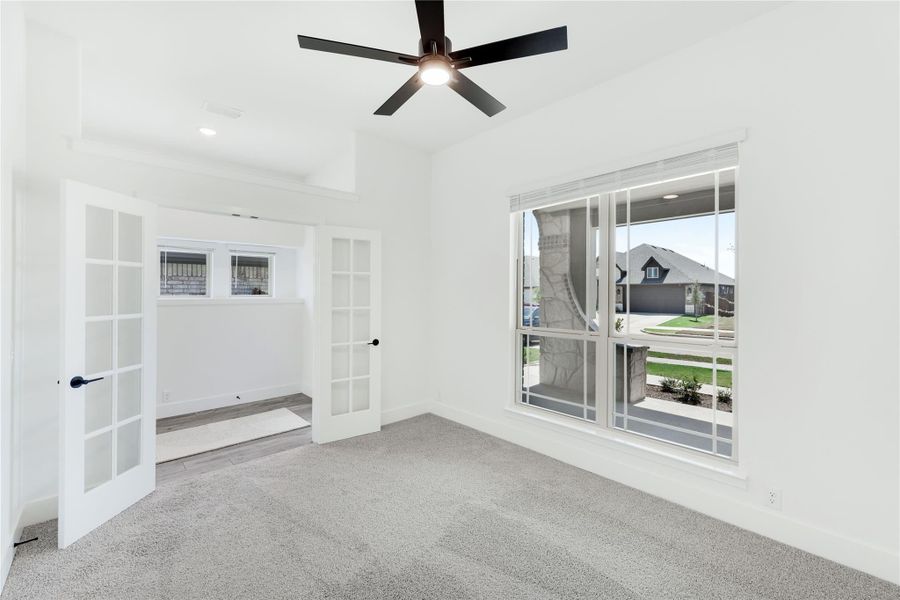 Empty bedroom with gray carpet, ceiling fan, large window, and French doors leading to an adjoining room Empty bedroom with gray carpet, ceiling fan, large window, and French doors leading to an adjoining room