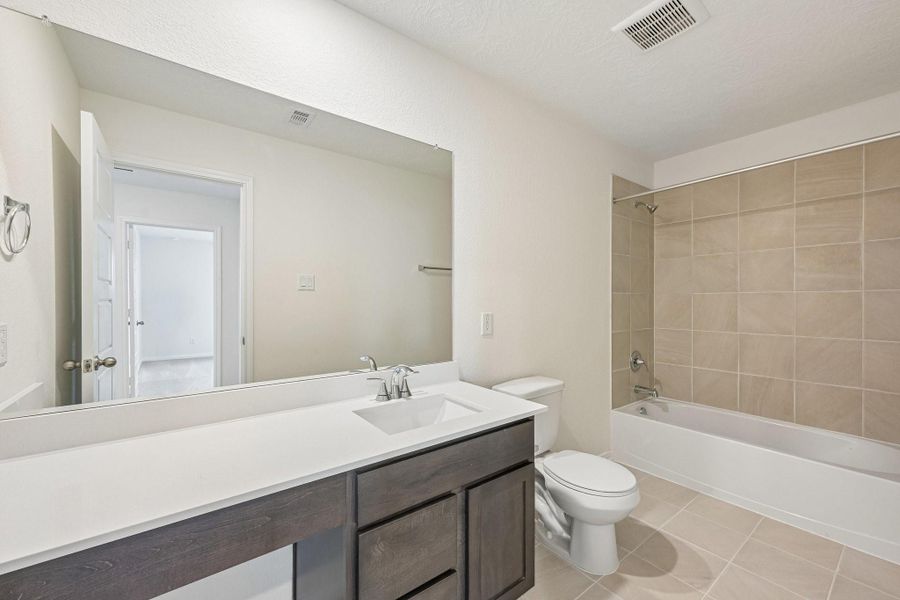 The secondary bathroom offers tile flooring, a bath/shower combination with a stylish tile surround, rich stained wood cabinetry with added knee space and elegant light countertops, a mirror, and sleek fixtures with modern finishes.