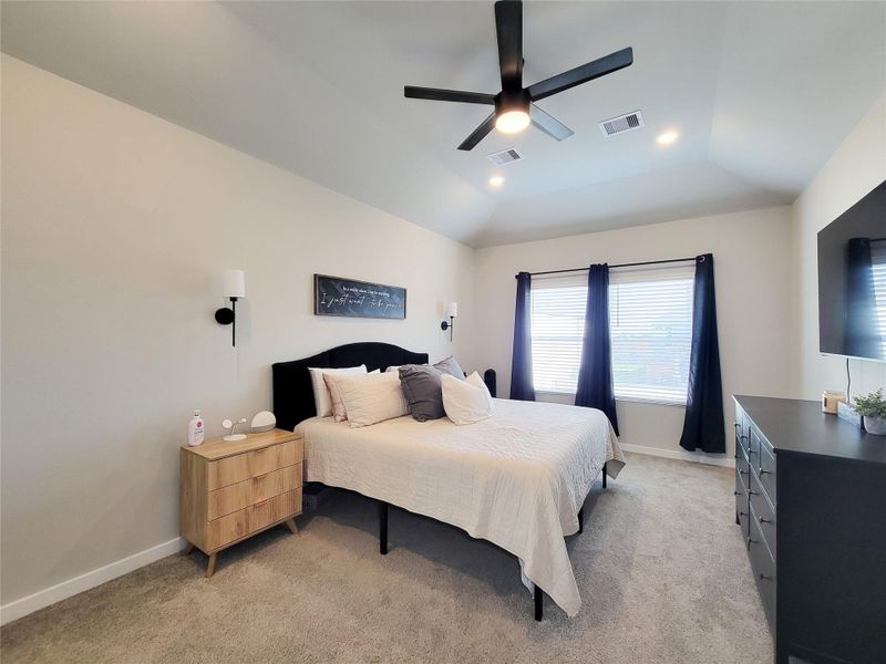 This Primary bedroom features a modern aesthetic with a vaulted ceiling and a sleek ceiling fan. It has a large window with navy curtains, allowing natural light to fill the space.