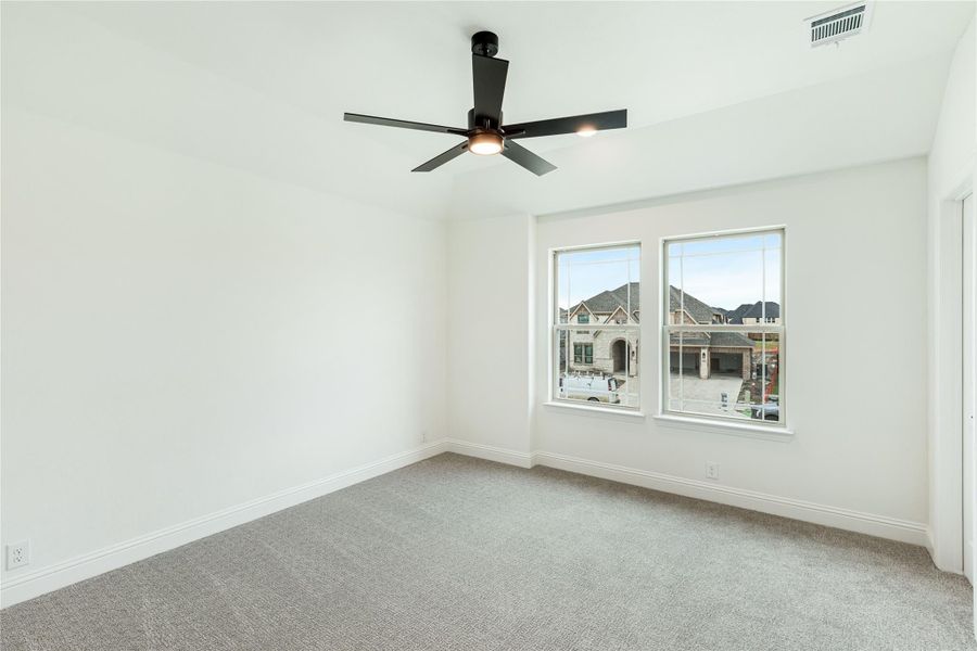 Empty bedroom with gray carpet, white walls, black ceiling fan, and two windows overlooking neighborhood homes.