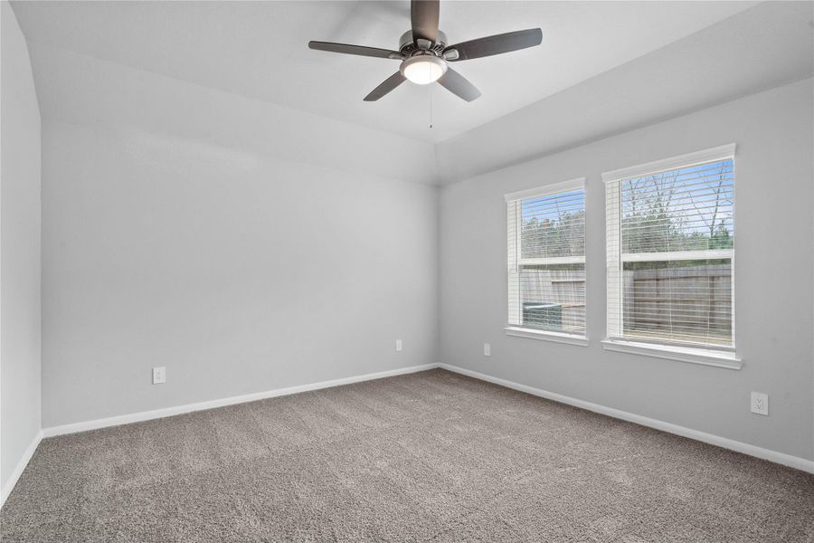 The primary bedroom provides a peaceful escape with a bright, neutral palette, sloped ceilings with ceiling fan, large windows (fitted with blinds), and plush carpet flooring, creating a calm and restorative atmosphere to end your day.