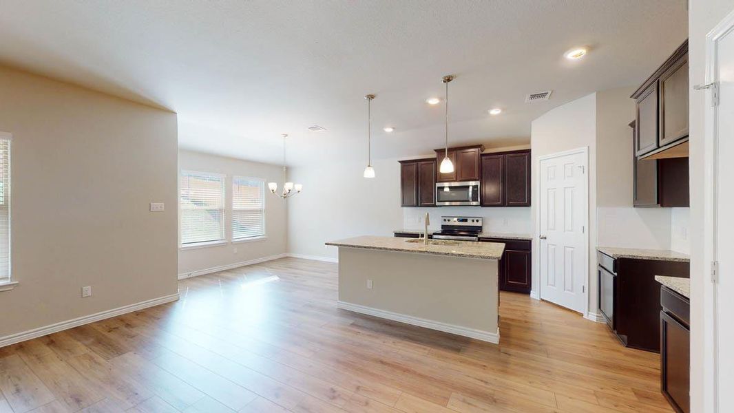 Kitchen with dark wood finish cabinets, hanging lights, light stone countertops, a kitchen island with sink, and stainless steel appliances