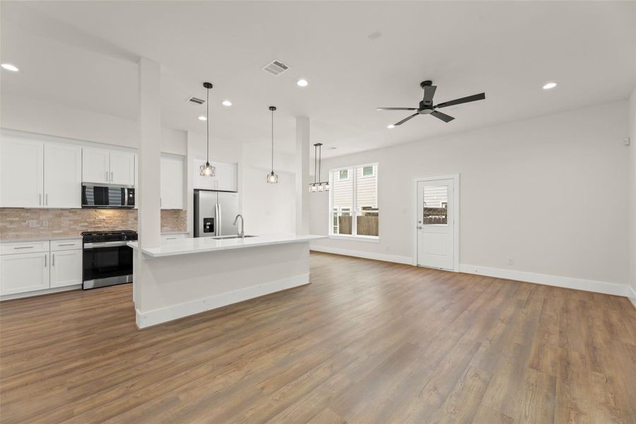 A spacious open-concept living area with a modern kitchen. The kitchen features white cabinetry, a large island, stainless steel appliances, and pendant lighting. The room is bright with wood like flooring, large windows, and a ceiling fan, creating a welcoming atmosphere.