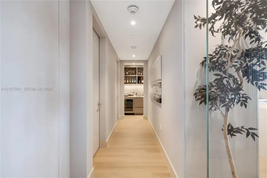 Hallway featuring light-toned wood-finish flooring, recessed lighting, and a glass-enclosed display area, leading to a built-in wet bar with cabinetry and stone-finish backsplash Hallway featuring light-toned wood-finish flooring, recessed lighting, and a glass-enclosed display area, leading to a built-in wet bar with cabinetry and stone-finish backsplash