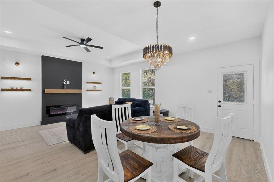 Open-concept living space featuring wood-finish flooring, a modern electric fireplace with a dark accent wall and light wood mantel, and illuminated floating shelves