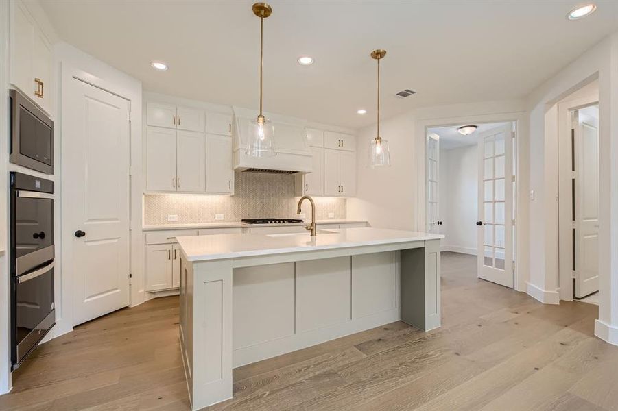 Kitchen with hanging light fixtures, light hardwood / wood-style floors, white cabinetry, an island with sink, and custom exhaust hood