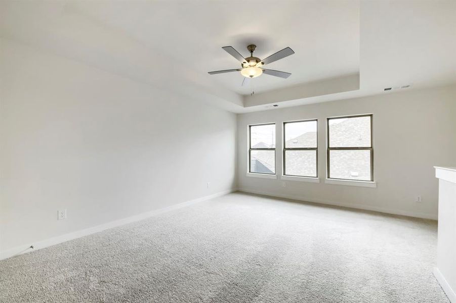 Spacious carpeted room featuring a ceiling fan with integrated lighting, a tray ceiling, and three large windows Spacious carpeted room featuring a ceiling fan with integrated lighting, a tray ceiling, and three large windows