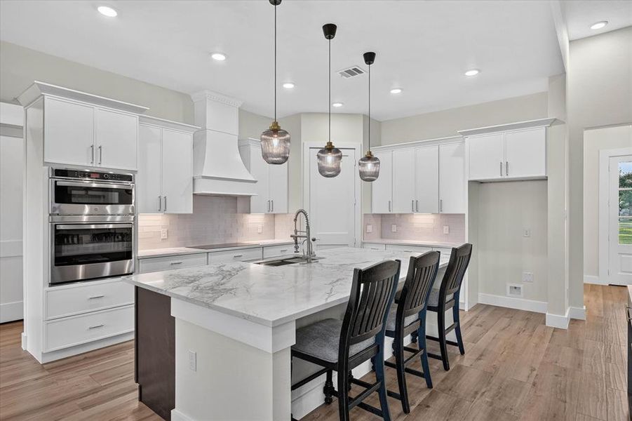 Gourmet kitchen featuring a large island with seating, stainless steel double ovens, white shaker cabinetry, a tile backsplash, and wood-finish flooring Gourmet kitchen featuring a large island with seating, stainless steel double ovens, white shaker cabinetry, a tile backsplash, and wood-finish flooring
