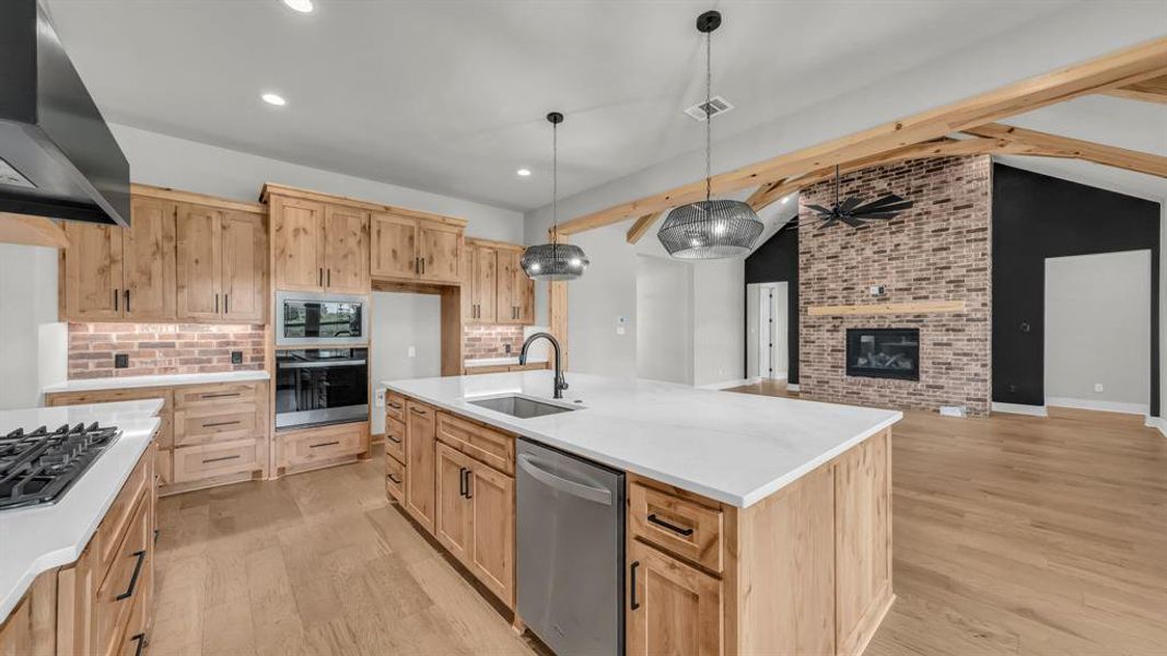 Open-concept kitchen and living area featuring wood-finish flooring, exposed wood beams, and a brick-finish fireplace