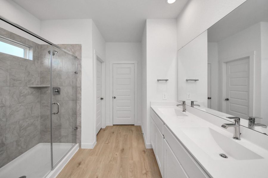 This modern primary bathroom features a spacious glass-enclosed shower with elegant tile work, a large double-sink vanity, and sleek fixtures, all set against a backdrop of bright, neutral tones and wood-look flooring.