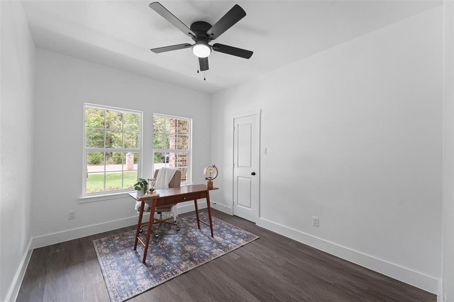Bright room featuring two large windows, dark wood-finish flooring, a modern black ceiling fan with integrated lighting, a white paneled door, and crisp white walls Bright room featuring two large windows, dark wood-finish flooring, a modern black ceiling fan with integrated lighting, a white paneled door, and crisp white walls