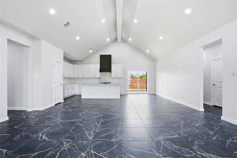 Expansive open-concept living space featuring vaulted ceilings with recessed lighting, large format dark tile flooring with white veining, a modern kitchen with white cabinetry and a contrasting black range hood, and sliding glass doors to the exterior