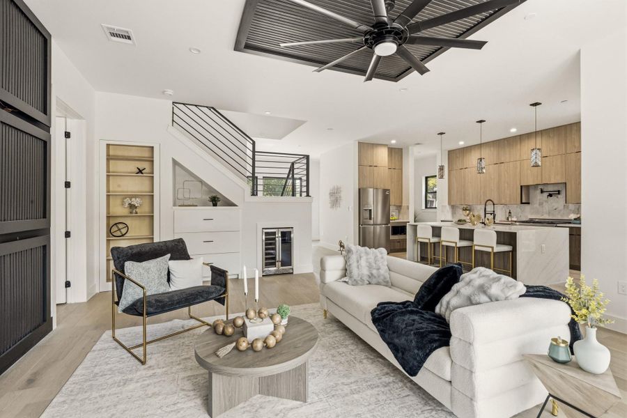 Open-concept living area featuring light wood-finish flooring, a modern kitchen with a large island, stainless steel appliances, and wood cabinetry, a staircase with black metal railings, and built-in under-stair shelving with a beverage cooler