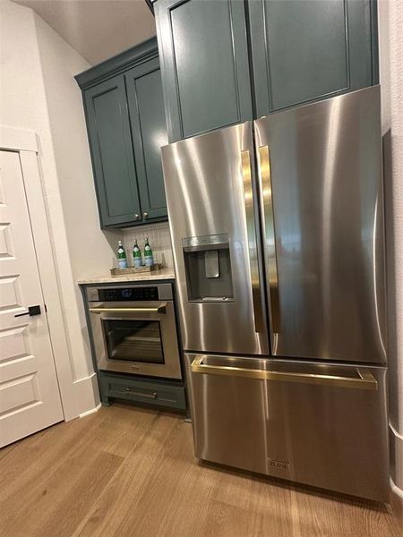 Kitchen featuring stainless steel appliances with brass-finish hardware, built-in oven, blue-toned cabinetry, light wood-finish flooring, and a white paneled door with black hardware