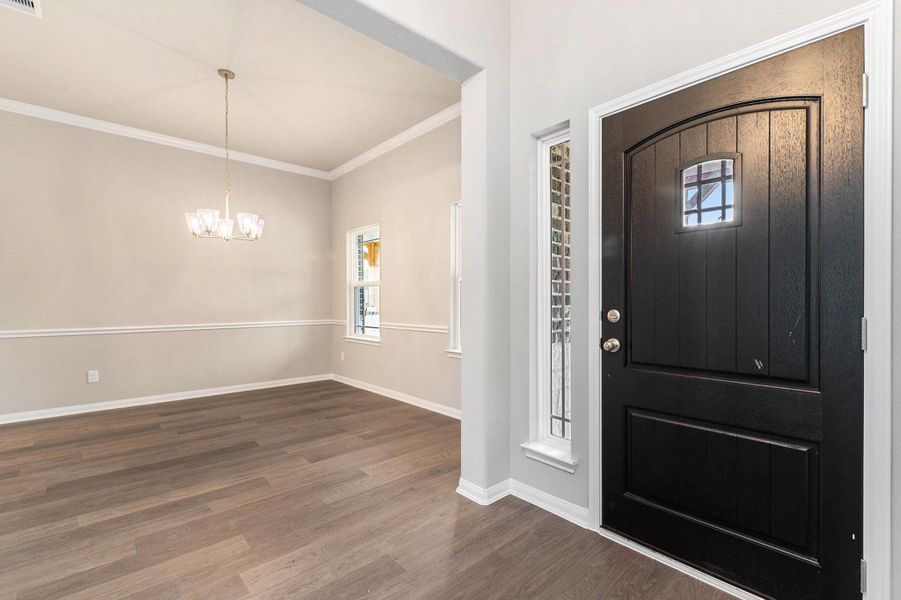 This welcoming entryway features a sleek black door with a small window, leading into a bright room with elegant wood flooring, neutral walls, and a stylish chandelier.