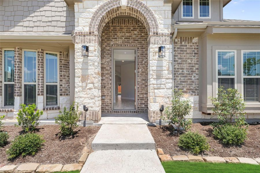 Home entrance with brick and stone arched doorway, wall sconces, and concrete walkway with landscaping