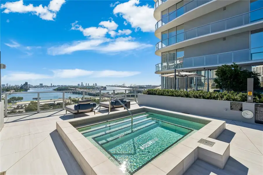 Terrace featuring a recessed square spa, large-format tile flooring, a glass panel railing, and a multi-story exterior with curved balconies