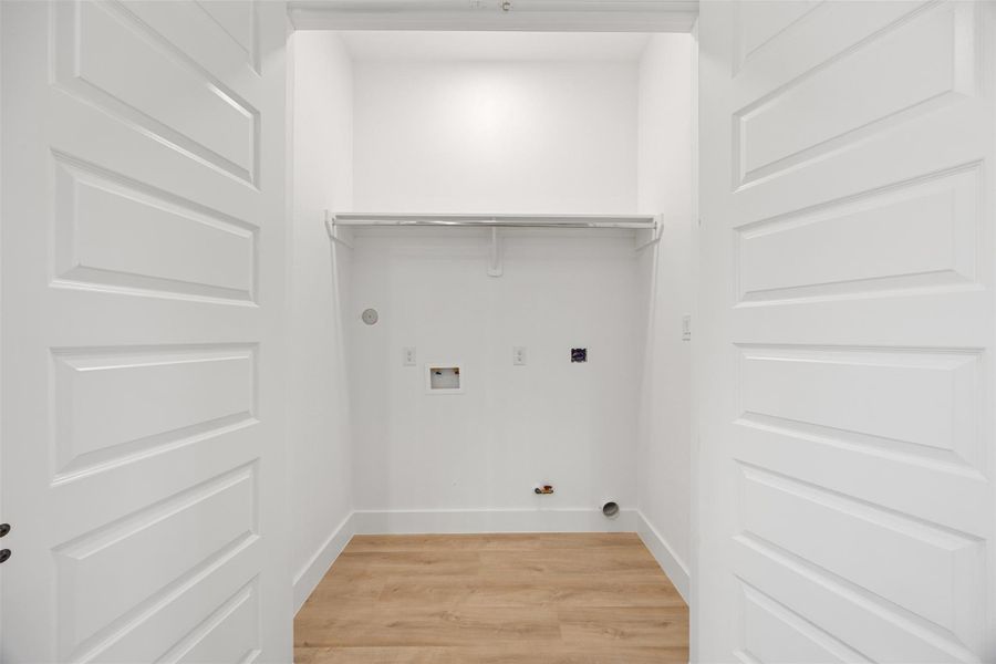 This photo shows a laundry area with hookups for a washer and dryer. It features white walls, a light wood-style floor, and a shelf above for storage. This photo shows a laundry area with hookups for a washer and dryer. It features white walls, a light wood-style floor, and a shelf above for storage.