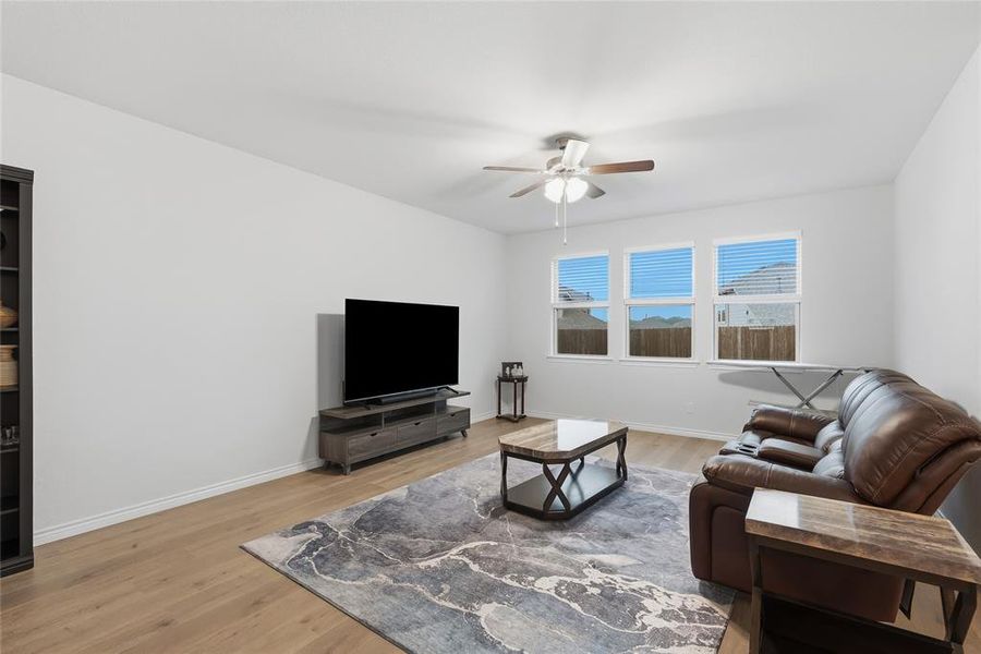 Spacious living area featuring light wood-finish flooring, white walls, and a ceiling fan with integrated lighting