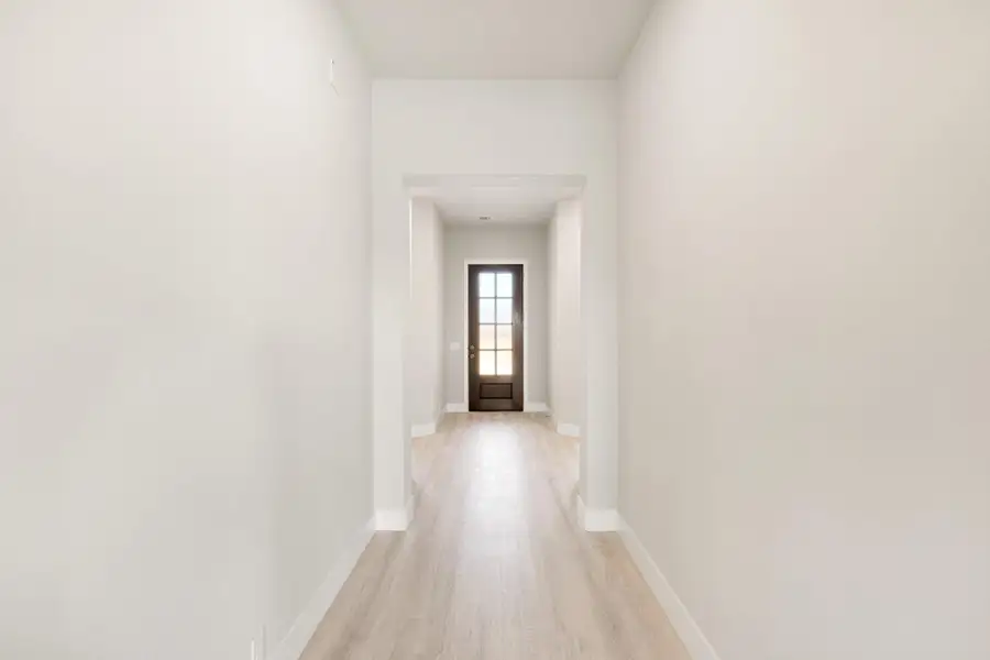 Narrow hallway with light wood floors, white walls, and dark front door with glass panels at the end