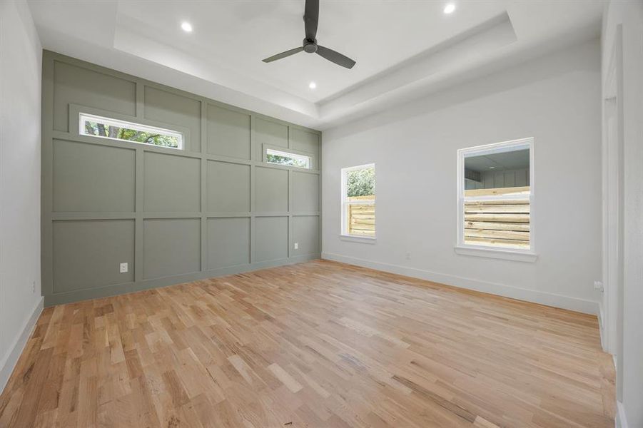 The primary bedroom offers a refined retreat with a decorative accent wall, ceiling fan, rich hardwood flooring, and an elegant tray ceiling detail that adds architectural character and sophistication. The primary bedroom offers a refined retreat with a decorative accent wall, ceiling fan, rich hardwood flooring, and an elegant tray ceiling detail that adds architectural character and sophistication.