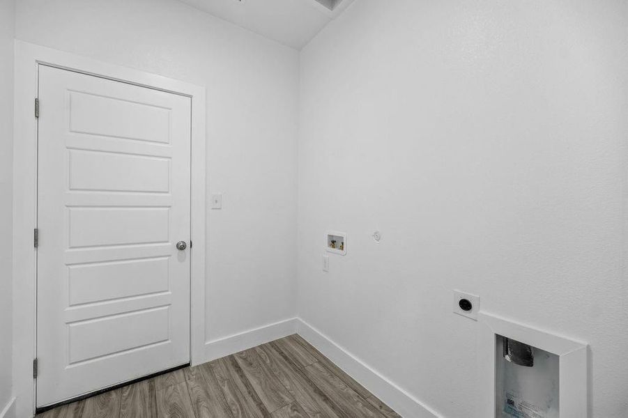 Utility room featuring wood-finish flooring, white baseboards, a white paneled door, and laundry hookups Utility room featuring wood-finish flooring, white baseboards, a white paneled door, and laundry hookups