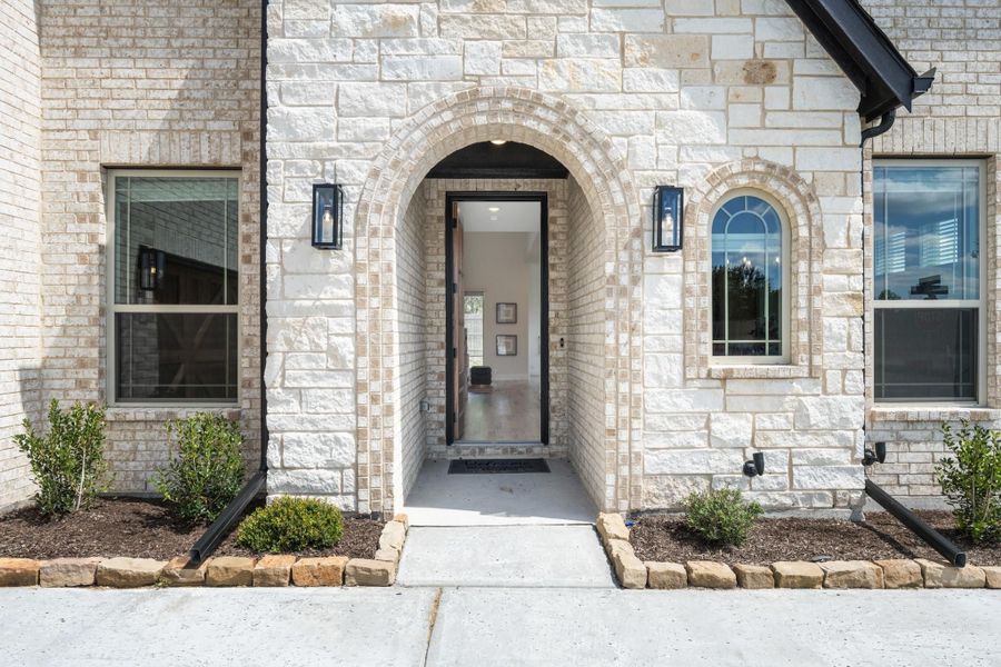 Home entrance with arched stone doorway, black door, and wall sconce lights on cream brick exterior