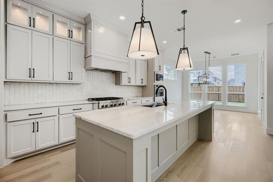 Spacious kitchen featuring an expansive island with a waterfall countertop, light-colored cabinetry with dark hardware, a herringbone pattern tile backsplash, and wood-finish flooring Spacious kitchen featuring an expansive island with a waterfall countertop, light-colored cabinetry with dark hardware, a herringbone pattern tile backsplash, and wood-finish flooring