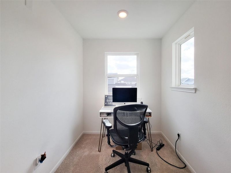 A compact, well-lit home office nook with a window view. It features neutral walls, carpet flooring, and space for a small desk and chair, ideal for focused work.
