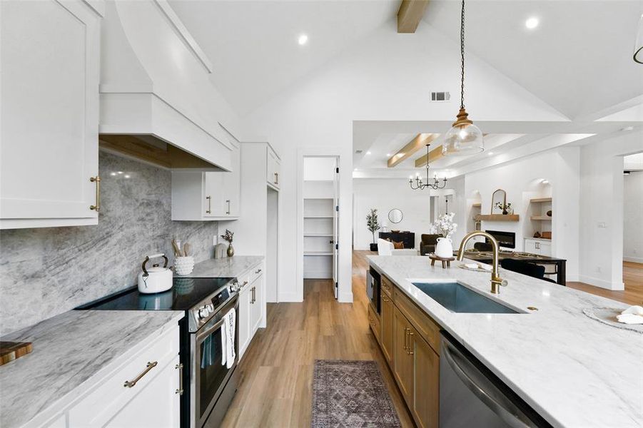 Open-concept kitchen featuring a spacious island with an undermount sink, stone countertops, light wood-finish flooring, white cabinetry, and a vaulted ceiling with exposed beams