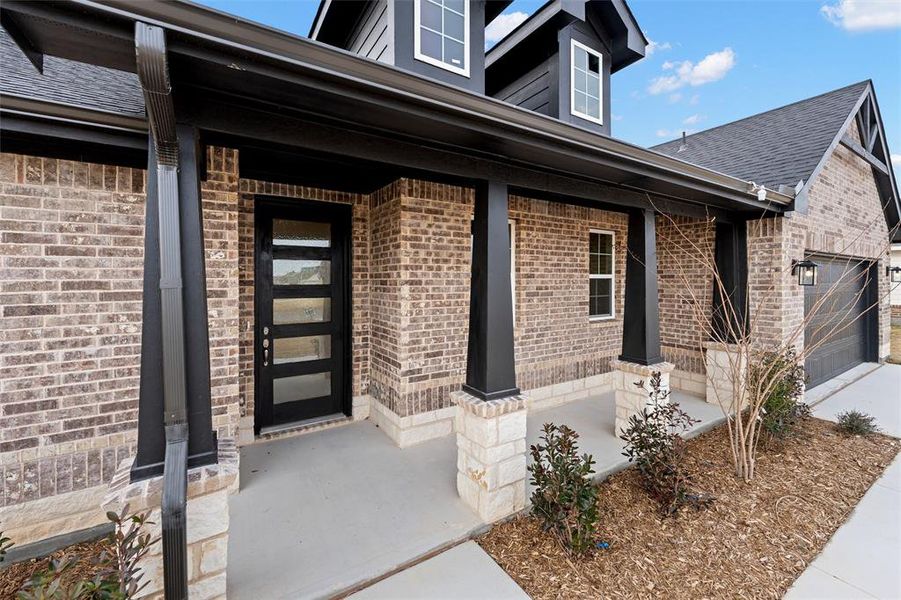 Entrance to property featuring a porch, brick siding, an attached garage, and roof with shingles