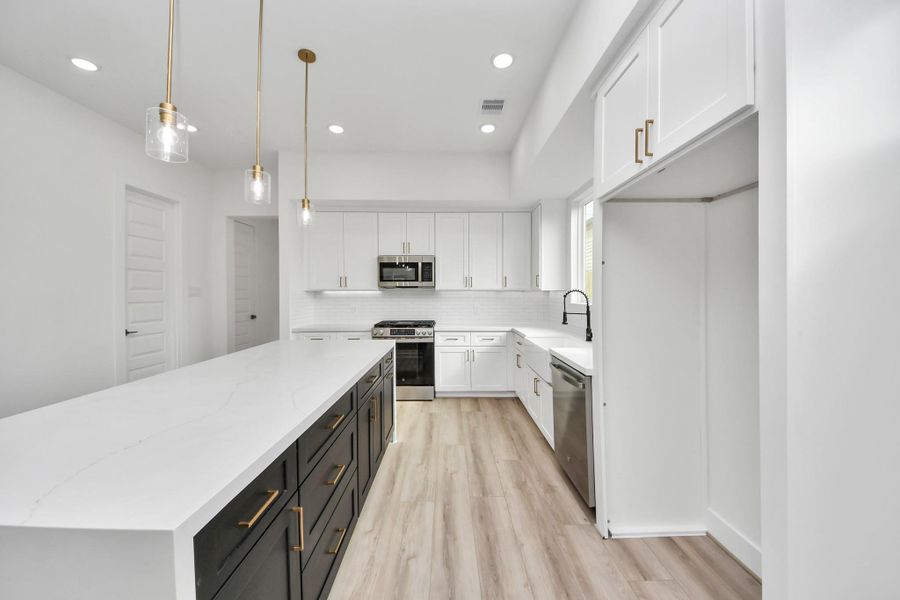 This modern kitchen features a spacious white island, sleek black and gold cabinetry, stainless steel appliances, and elegant pendant lighting, all set against light wood flooring for a bright, contemporary look.