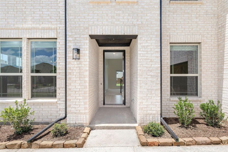 Front entry of home with light brick exterior, black-framed door, wall lantern, and small landscaped shrubs