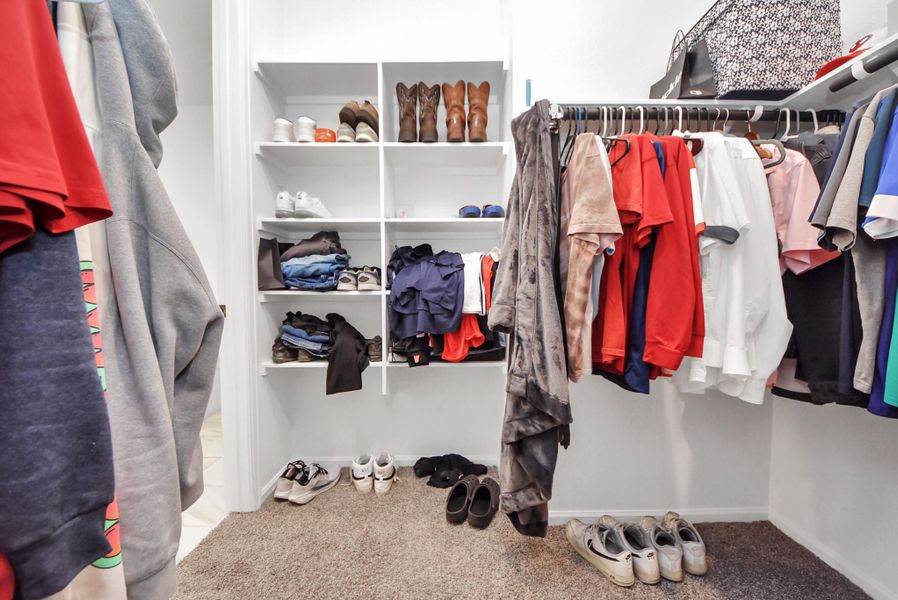 This photo shows a spacious walk-in closet with carpeted flooring. It features multiple shelves and hanging rods for organized clothing and shoe storage. The closet is well-lit and efficiently designed for easy access.