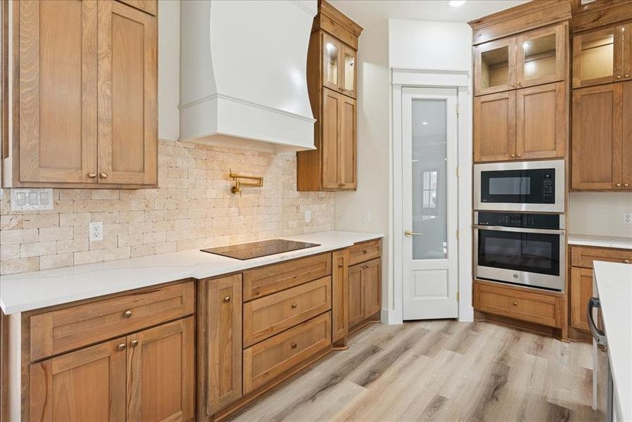 Kitchen with brown cabinetry, stainless steel appliances, glass insert cabinets, and decorative backsplash