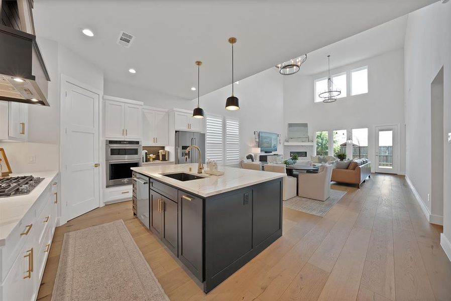 Spacious kitchen featuring an island with a sink, white cabinetry, integrated stainless steel appliances, and wood-finish flooring extending into a high-ceiling living area with large windows Spacious kitchen featuring an island with a sink, white cabinetry, integrated stainless steel appliances, and wood-finish flooring extending into a high-ceiling living area with large windows