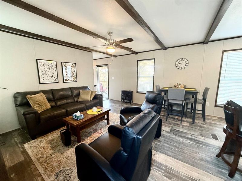 Living room featuring beam ceiling, wood finished floors, ceiling fan, and a wood stove