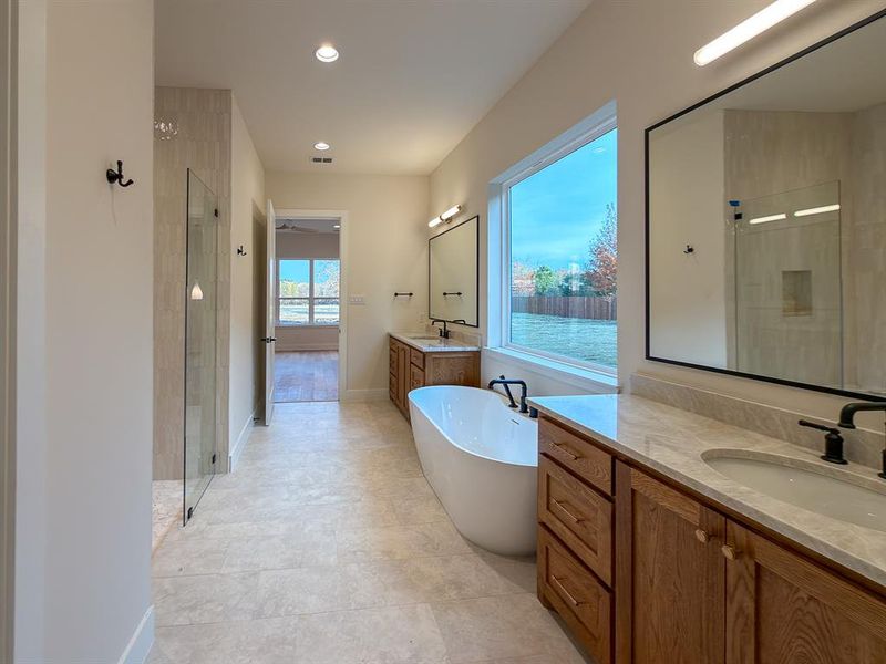 Spacious bathroom featuring a freestanding soaking tub, dual wood-finish vanities with light stone countertops, a large window, and a walk-in glass shower